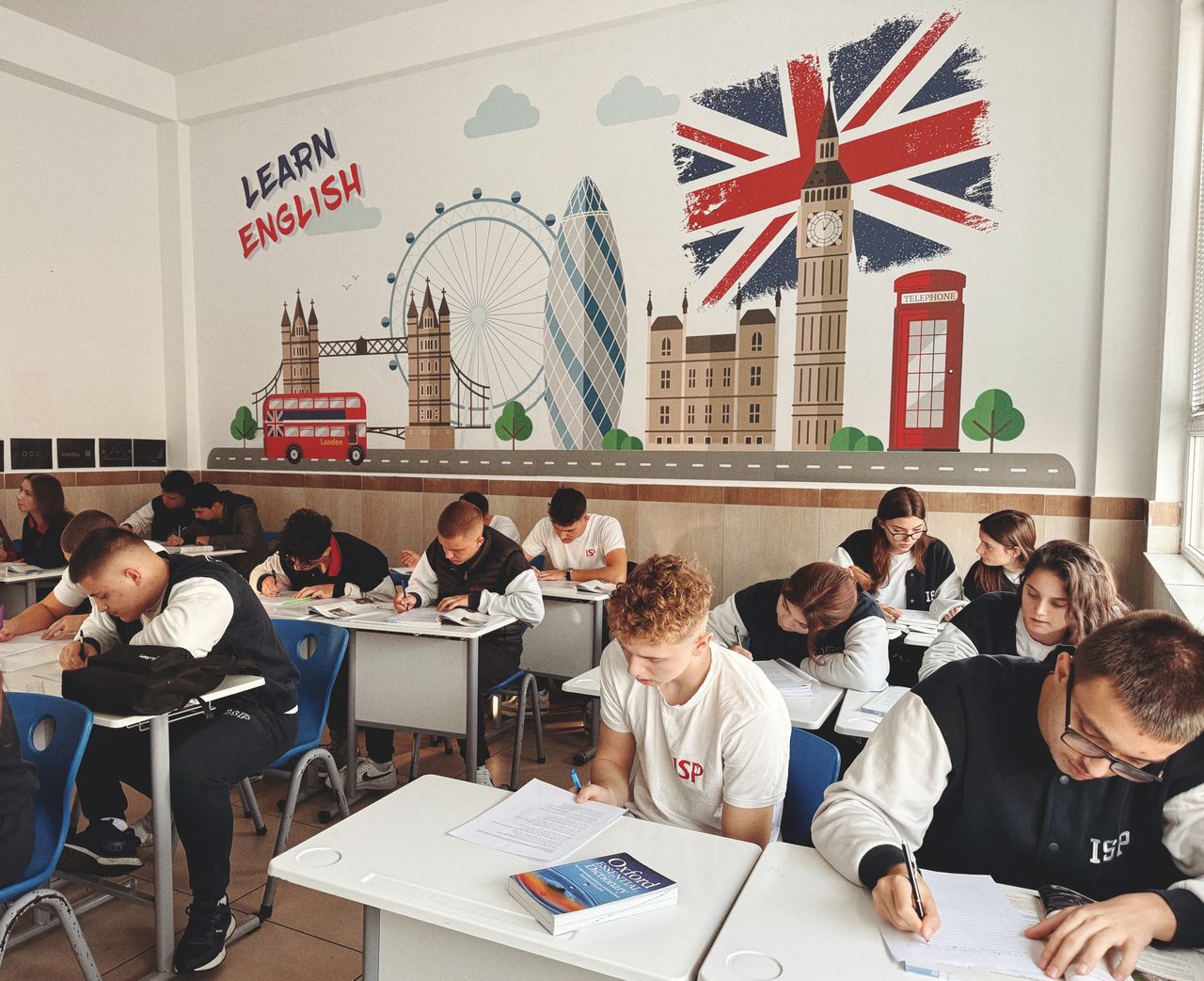 About Students studying English in a classroom with British themed decor and mural.